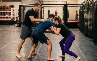 MMA coach guiding two adult students through clinch-to-ground technique at Arizona Combat Sports, a long-established MMA gym in Arizona
